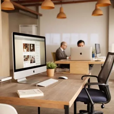 Modernes Büro mit Holzschreibtischen, großen iMac-Computern im Fordergrund, dem ein Mitarbeiter und deshalb Know-how feht. Dahinter zwei Personen, die an Computerns arbeiten.