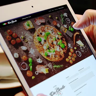 Eine Person hält ein Tablet in der Hand, auf dem die responsive Website eines Restaurants mit einer großen Pizza abgebildet ist, die von Vorspeisen, Beilagen und einem Online-Formular umgeben ist - ein Beispiel für gutes Design, das sich perfekt mit Wordpress realisert wurde.