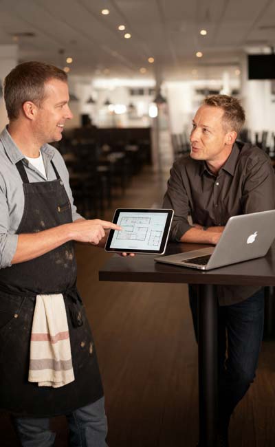 Zwei Männer unterhalten sich an einem Stehtisch in einem modernen Restaurant; der eine in einer Schürze hält ein Tablet mit IT Planungen in der Hand, der andere lehnt am Tisch neben einem geöffneten Laptop und bespricht die Möglichkeiten einer kostenlosen Beratung.