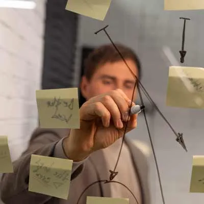 Eine Person im Anzug zeichnet mit einem Marker Diagramme auf eine Glastafel, umgeben von Klebezetteln, auf denen Marketing Startegie und Werbekonzepte skizziert werden.