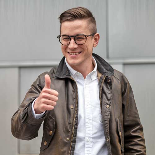 Ein lächelnder Projektentwickler mit Brille, brauner Jacke und weißem Hemd zeigt den Daumen nach oben, während er im Freien vor einem grauen Hintergrund steht und sich über eine moderne Website freut.
