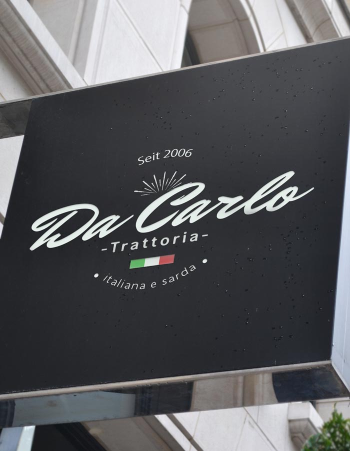 Beispiel Schild, ein schwarzes Restaurantschild mit der Aufschrift "Seit 2006 Da Carlo Trattoria italiana e sarda" und einer kleinen italienischen Flagge unterhalb des Textes ist an einer Hauswand angebracht.