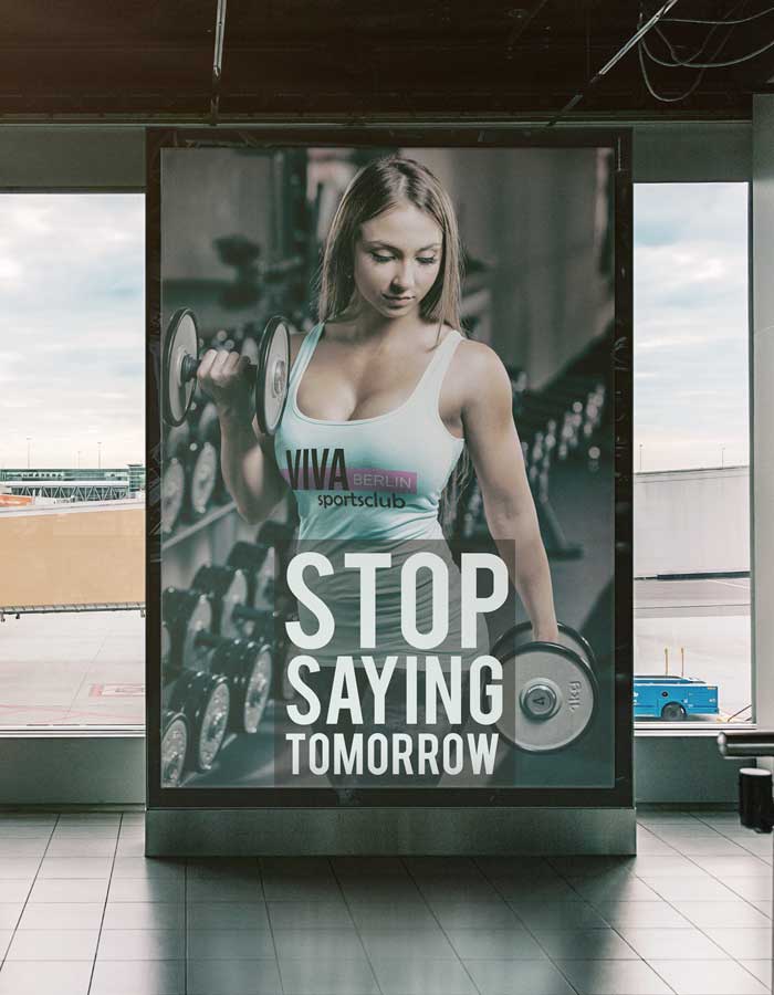 Ein Plakat in einem Fitnessstudio zeigt eine Frau, die Hanteln stemmt, mit dem Text "STOP SAYING TOMORROW" und "Viva Berlin Sportsclub". Fitnessgeräte, ein Fensterblick und professionelle Werbedrucke schaffen eine inspirierende Atmosphäre.
