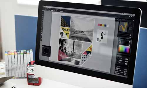 Auf einem Computermonitor wird ein Grafikdesignprojekt mit Fotos, Formen und Farbpaletten angezeigt, das in einer Druckdesignsoftware geöffnet ist. Im Hintergrund sind verschiedene Büromaterialien zu sehen.