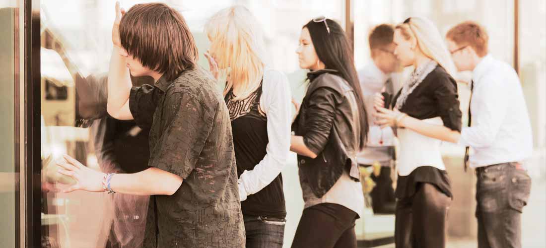 Eine Gruppe von Menschen steht in einer Schlange vor einem Gebäude, einige blicken durch ein Fenster. Sie sind in Freizeit- und Geschäftskleidung gekleidet und scheinen zu warten oder zu beobachten - vielleicht, weil sie sich bemühen, die Kundenzufriedenheit im Inneren zu steigern.