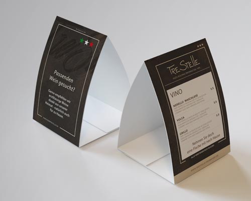 Zwei dreieckige Tischaufsteller stehen auf einer weißen Fläche. Der linke Aufsteller ist mit deutschem Text und Sternen versehen, während der rechte eine italienische Weinkarte unter der Überschrift Tre Stelle präsentiert.