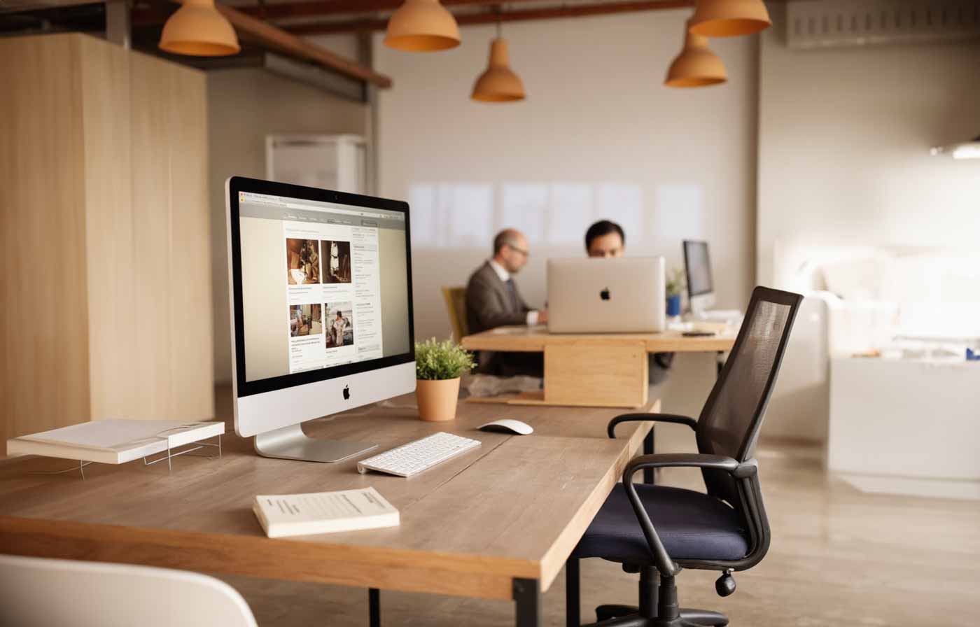 Modernes Büro mit Holzschreibtischen, großen iMac-Computern im Fordergrund, dem ein Mitarbeiter und deshalb Know-how feht. Dahinter zwei Personen, die an Computerns arbeiten.