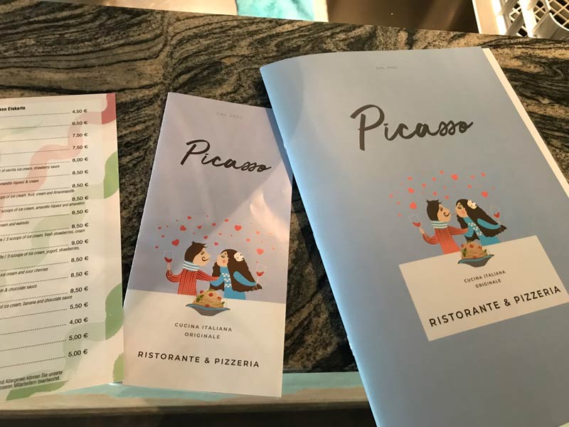 Auf einem Marmortisch neben einer Preisliste liegen zwei Speisekarten des „Picasso Ristorante & Pizzeria“. Auf den Speisekarten ist ein Paar abgebildet, das sich unter herabfallenden Herzen eine Portion Pasta teilt.