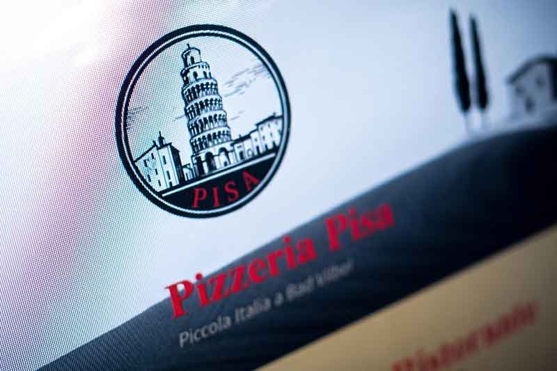 Nahaufnahme eines Computerbildschirms, auf dem das Logo der „Pizzeria Pisa“ zu sehen ist, das den Schiefen Turm von Pisa, umrissene Gebäude, zwei Bäume sowie das Wort „PISA“ in Grün und Rot zeigt.