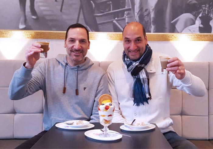 Zwei Männer sitzen an einem Tisch, lächeln und heben ihre Kaffeetassen. Zwischen ihnen steht ein Dessert mit Obst und Waffeln auf dem Tisch. Der Schauplatz scheint ein Café mit heller Einrichtung zu sein.