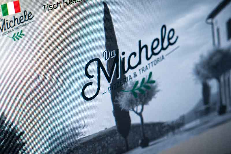 Nahaufnahme eines Computerbildschirms, auf dem eine responsive Website der „Da Michele Pizzeria & Trattoria“ zu sehen ist, die in den Farben der italienischen Flagge gehalten ist und im Hintergrund das Bild einer Zypresse und einer Steinmauer zeigt – was das authentische kulinarische Erlebnis perfekt widerspiegelt.