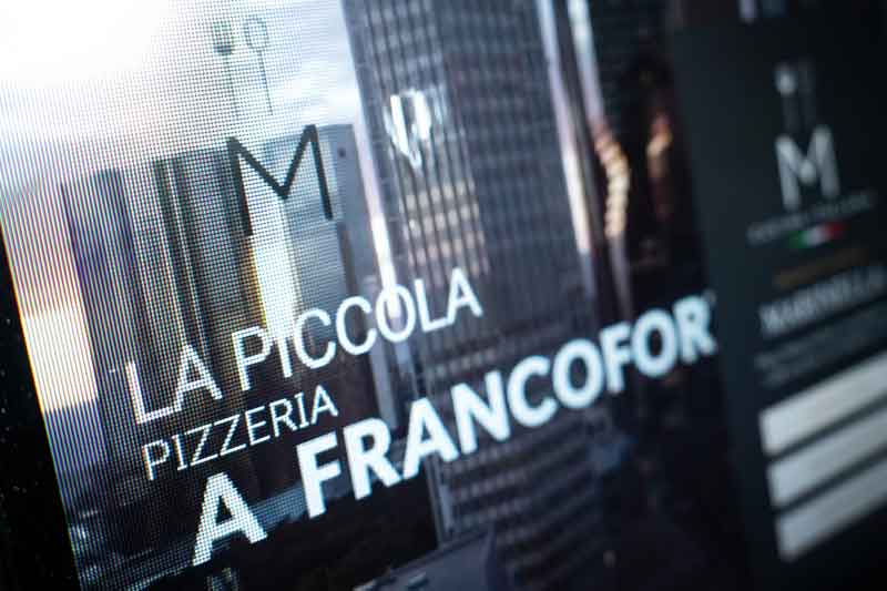 Auf einer digitalen Restauranttafel steht „La Piccola Pizzeria a Francofor“, wobei sich die Gebäude der Stadt im Glas hinter dem Text spiegeln – ein moderner Akzent, der die Gastronomieszene hervorhebt und sowohl vor Ort als auch auf den Webseiten auf das Lokal aufmerksam macht.