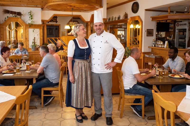 In einem gut besuchten Restaurant steht ein lächelnder Koch in Uniform neben einem „Kunde“ in traditioneller Tracht, während die Gäste an Holztischen in einem warm beleuchteten, rustikalen Ambiente Speisen und Getränke genießen.
